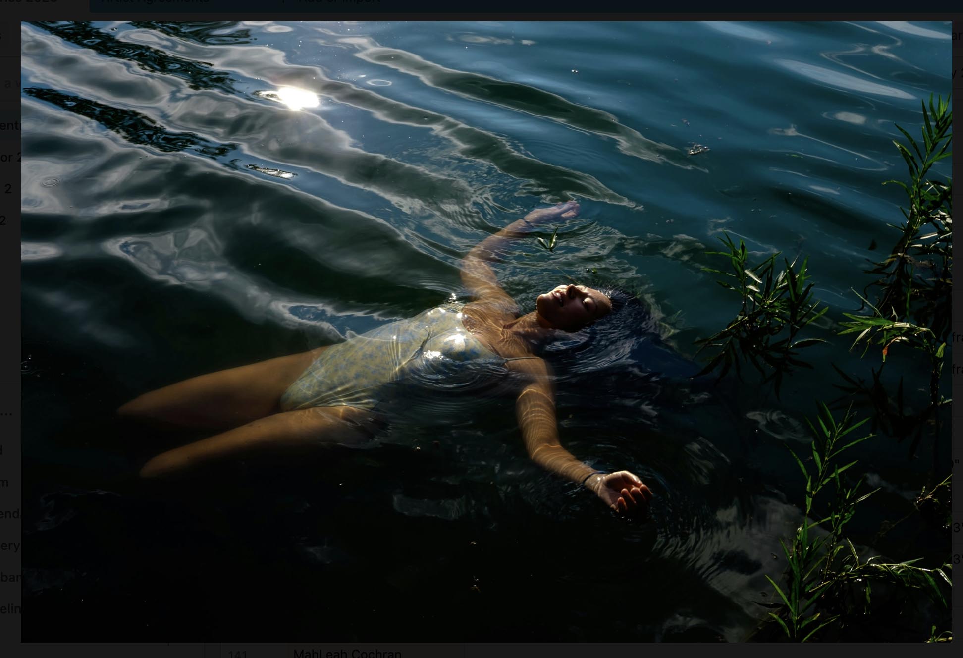 A light-skinned person in a white body suit, floating in a body of water with the sun reflecting in the water. Pine branches crowd the right side of the piece.