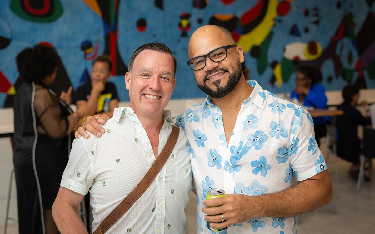 Two people, both with short haircuts and button-down shirts, smile at the camera in front of a large, multi-colored mural. The person on the left is white and is wearing a satchel. The person on the right is black and is wearing dark glasses. The second individual is wearing a visible wedding ring. They have their arms around each other's shoulders in a familiar pose