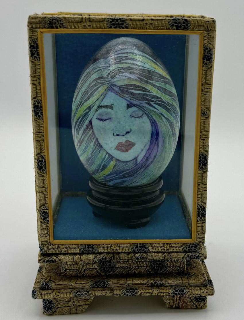 A tan woven box with black detailing and a dark blue interior. Inside of it is the face of woman with closed eyes and a somber expression. The woman has a light blue complexation and blue/green colored strands of hair that frame her face ; her cheeks, eyes and lips are slightly rosy.