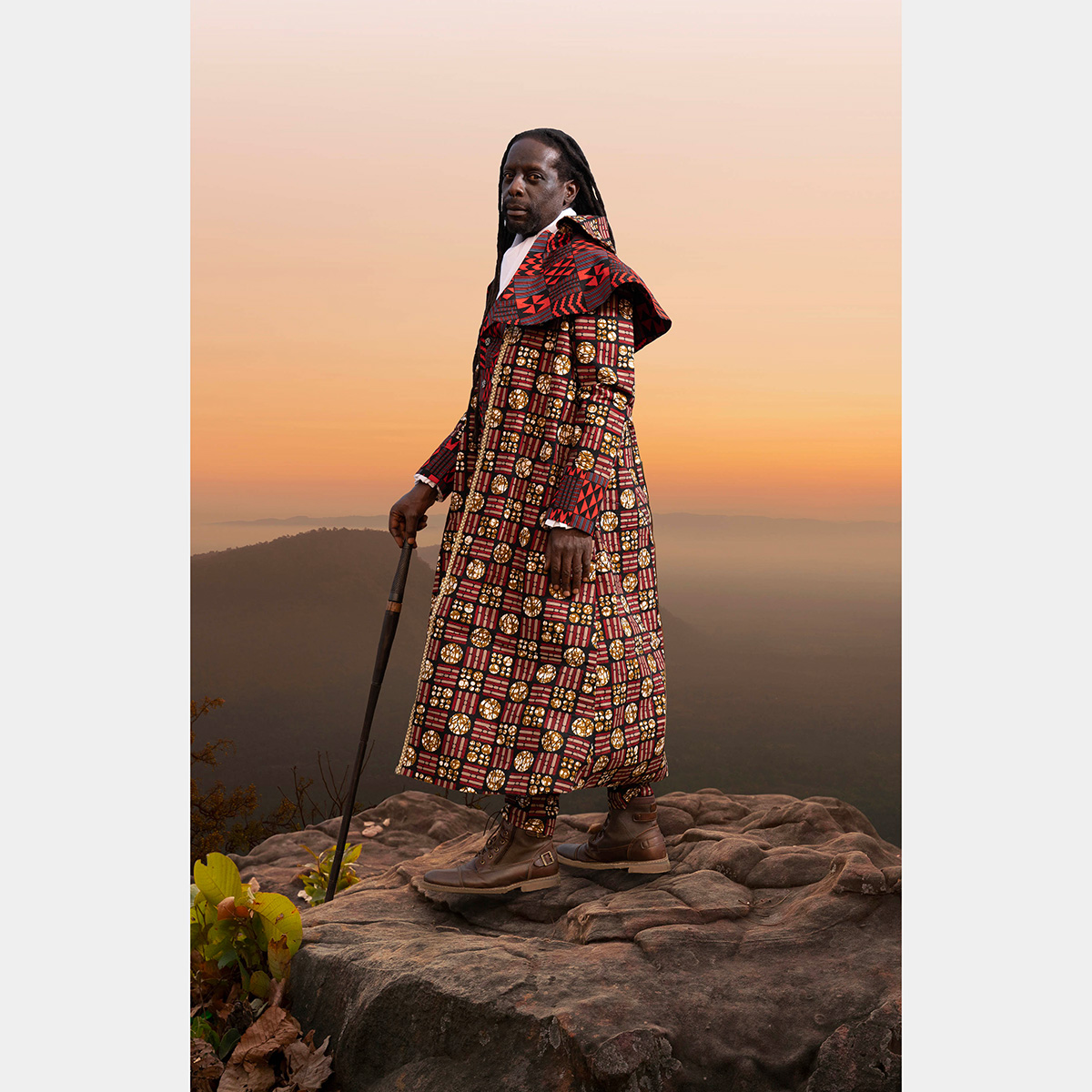 A Black man wears a red robe and stands on top of a mountain at sunset