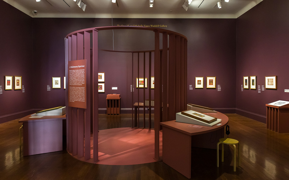 Circular structure made of vertical slats sits in the middle of an exhibition space surrounded by tabletop displays and framed artwork hanging on walls.