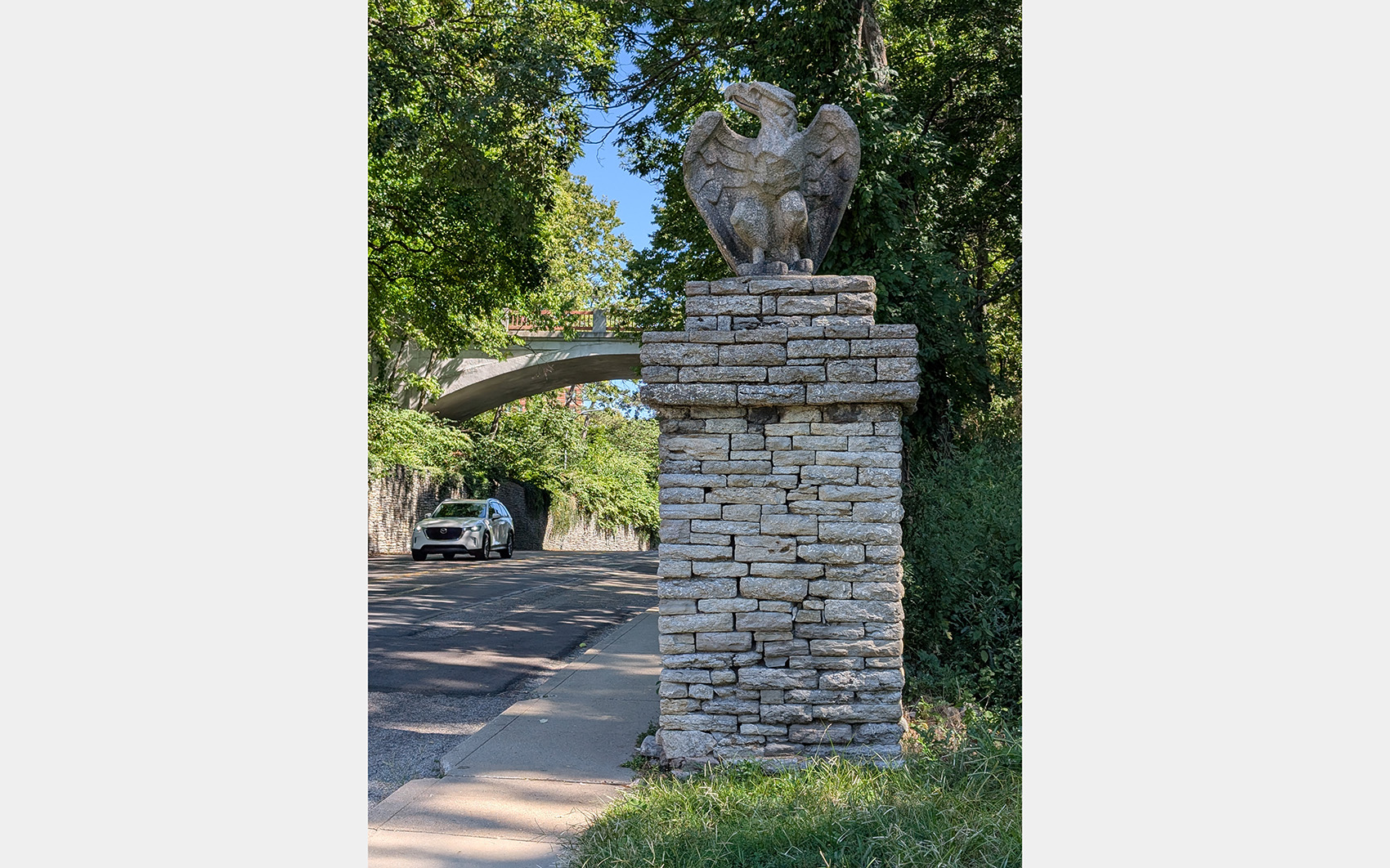 Stone eagle sculpture perched on stone masonry pedestal next to a road with trees and bridge in background.