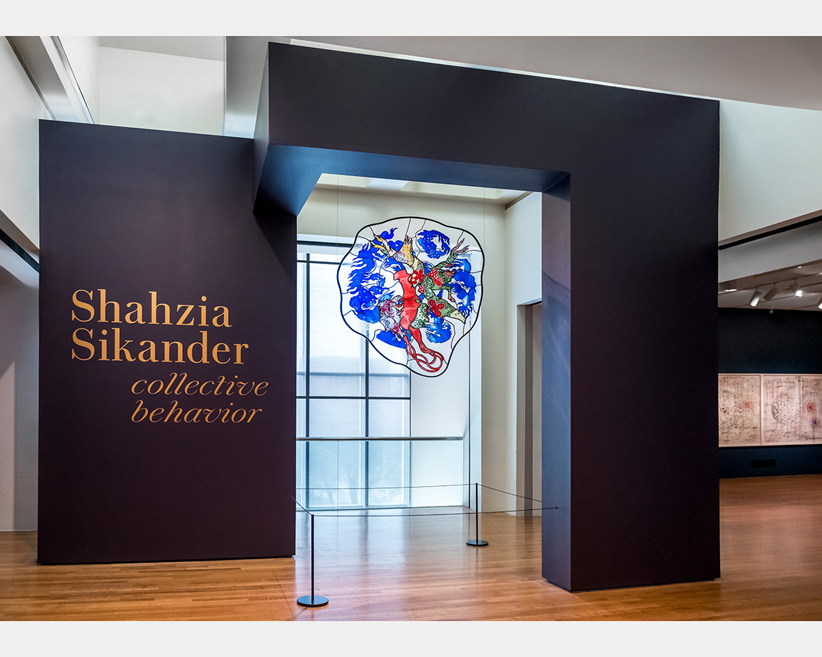 A back-lit, stained-glass artwork hangs from a ceiling and is framed by purple walls inside a gallery space. Gold lettering one of the walls states "Shahzia Sikander collective behavior".