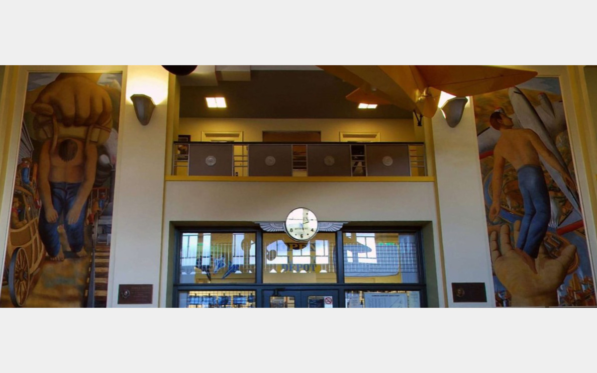 Interior of 2-story building lobby with balcony with a clock in the center and two murals on either side.