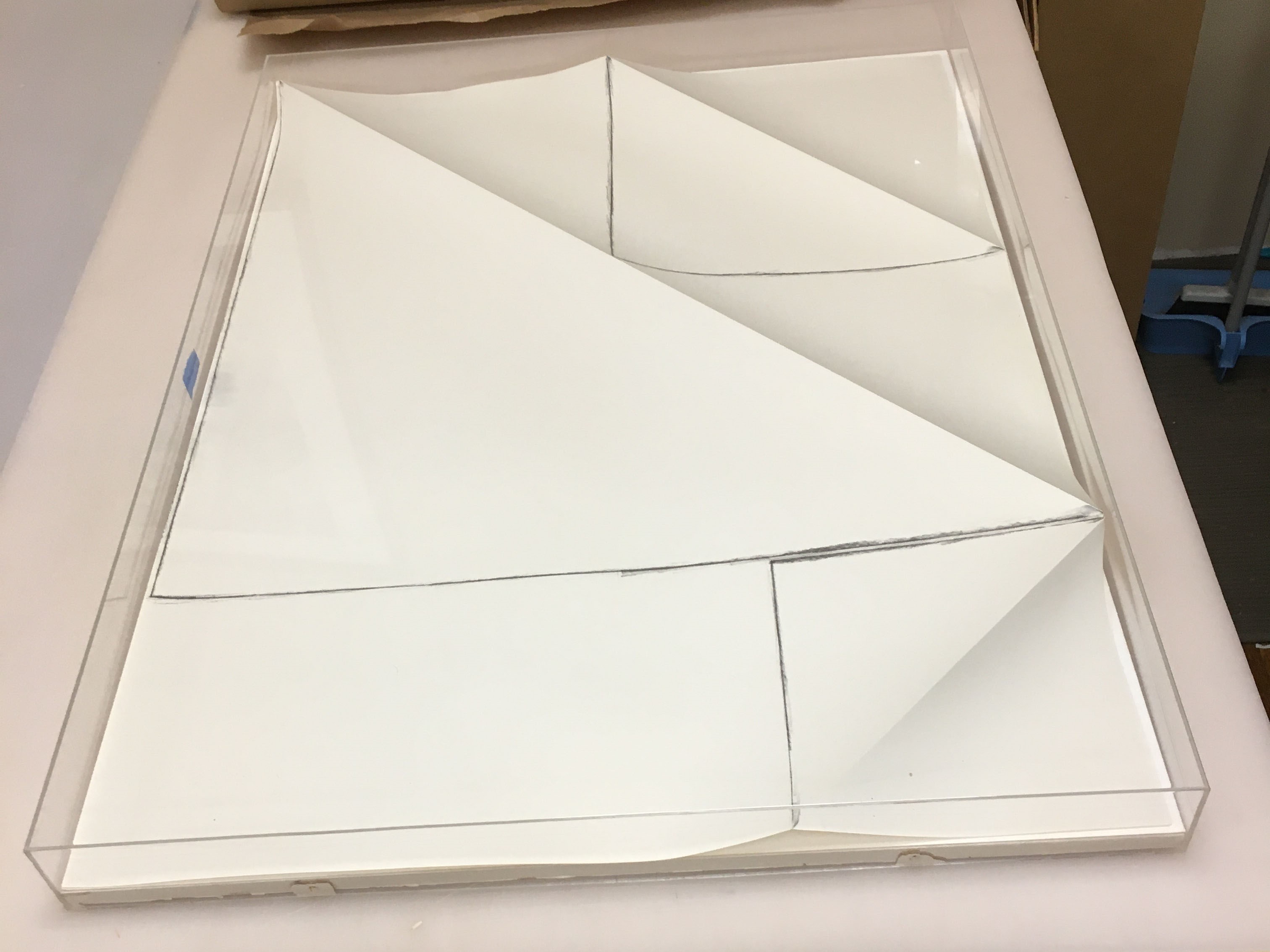 Folded paper artwork with linear black crayon markings mounted inside a transparent acrylic case.