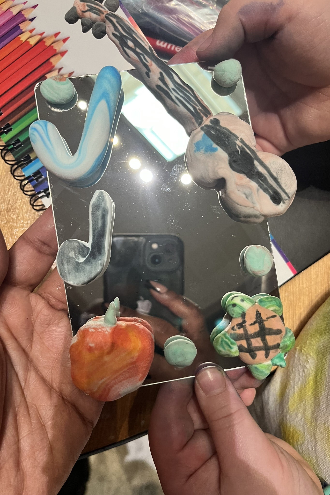 View from above of three hands holding a piece of artwork of figurines mounted onto a small mirror.