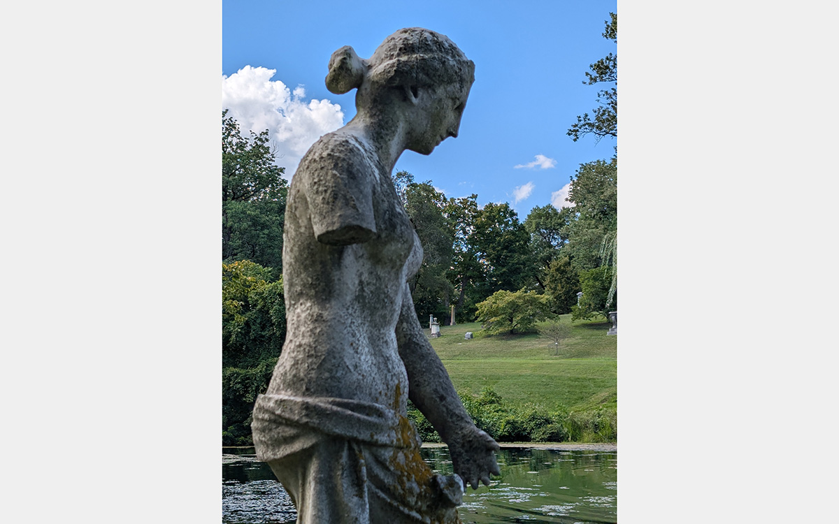 A marble sculpture of a woman wearing drapery from the waist down and nude from the waste up. A green landscape with trees, pond and ,blue sky is in the background.