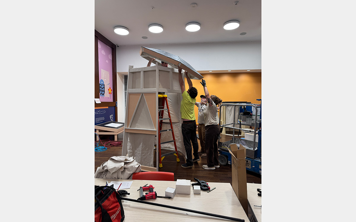 Three people lift a roof onto a wood structure inside a large room.