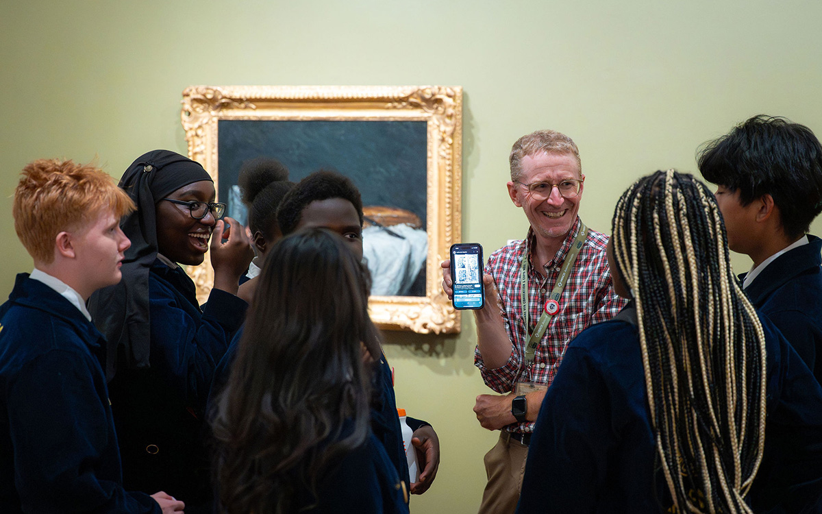 A man holds up a smartphone showing the screen to a group of students gathered around a painting.