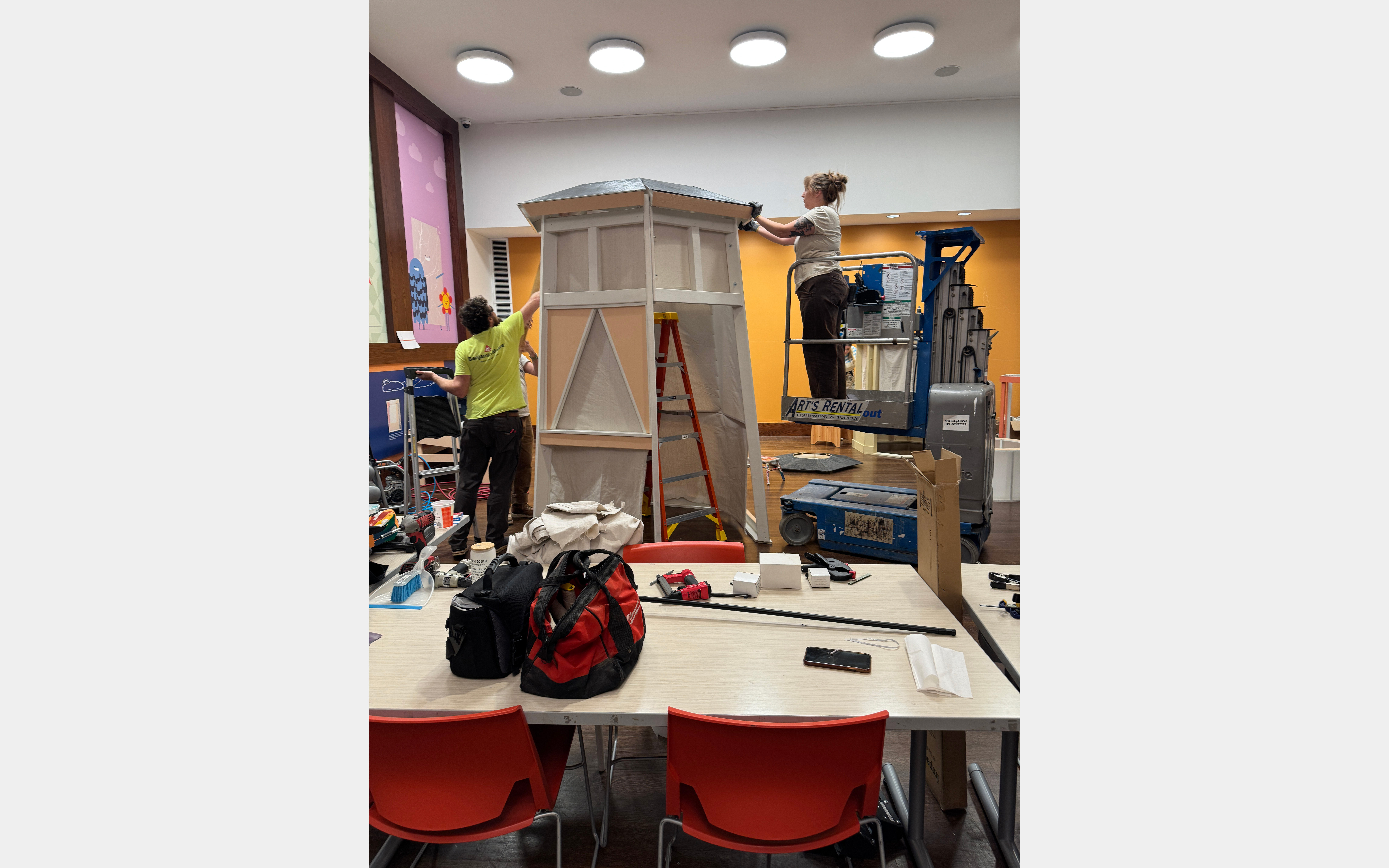 Two people building a tower-like structure inside a large room. One person works from a mechanized lift.